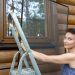 Young adult beautiful slim sporty caucasian woman stand on metal ladder and installing window mosquito insect fly screen protection. Female person housewife attach plastic mesh home wall summer day.