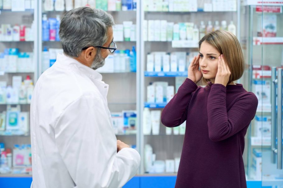 Side view of female customer grabbing head while male chemist wearing white lab coat and eyeglasses helping, offering medicaments from headache. Young woman consulting with pharmacist in drugstore.