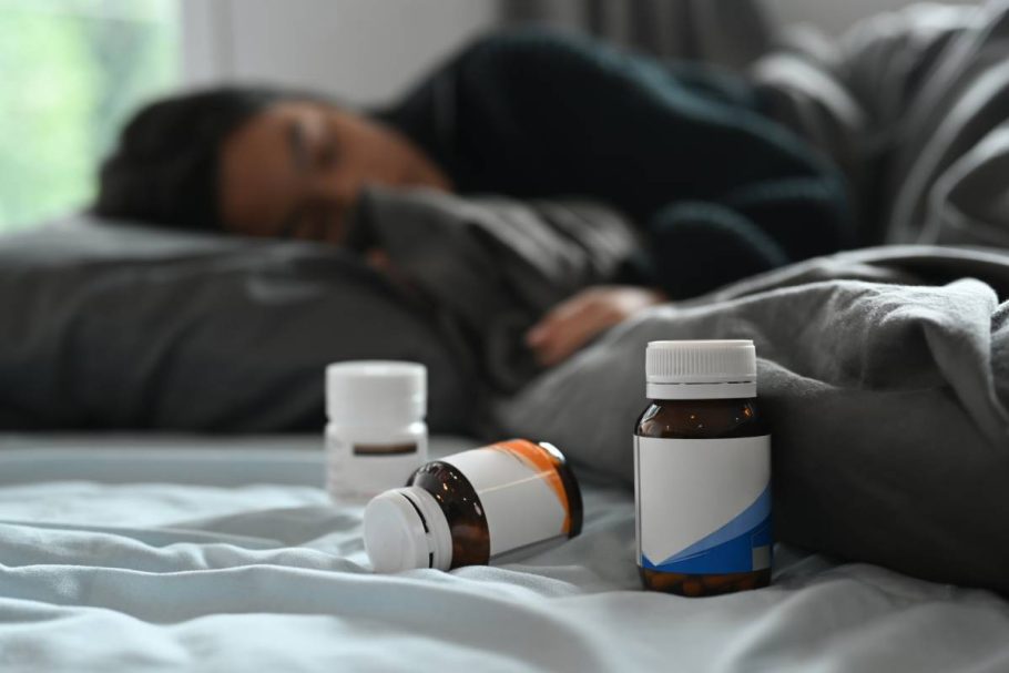 Selective focus on pill bottles and ill little girl lying in bed under blanket with high fever.