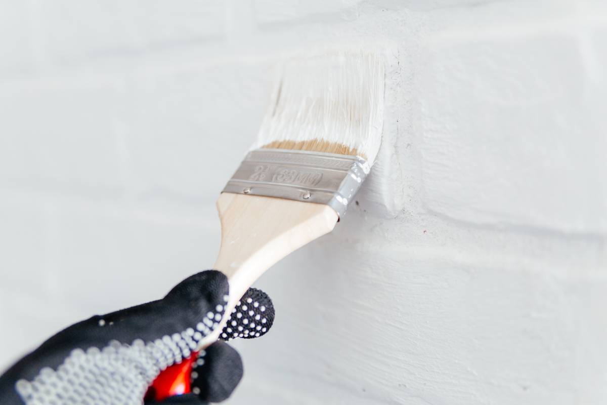 Close-up of a worker's hand with a brush painting a brick wall with white paint.