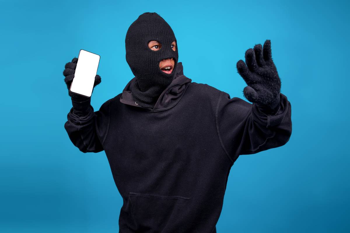 Portrait Of Funny Robber Wearing Balaclava Defending While Showing Blank Smartphone Screen Over Blue Background